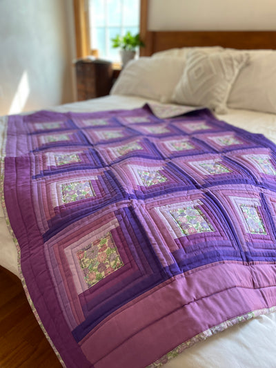 This heirloom lap quilt with a log cabin pattern with shades of lavenders and purples and a small print floral cover the front of this quilt. The binding and backing match the center floral print. It is made with a warm, heavy weight cotton/wool blend batting inside.