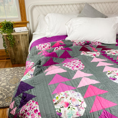 A handmade, heirloom, patchwork of pink and magenta flying geese on a grey/gray background with floral prints, watercolor bird fabric, magenta backing, straight line top quilting, machine top stitched binding, on this large king size quilt