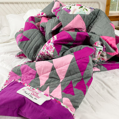 A bunched up handmade, heirloom, patchwork of pink and magenta flying geese on a grey/gray background with floral prints, watercolor bird fabric, magenta backing, straight line top quilting, machine top stitched binding, on this sweet queen size quilt