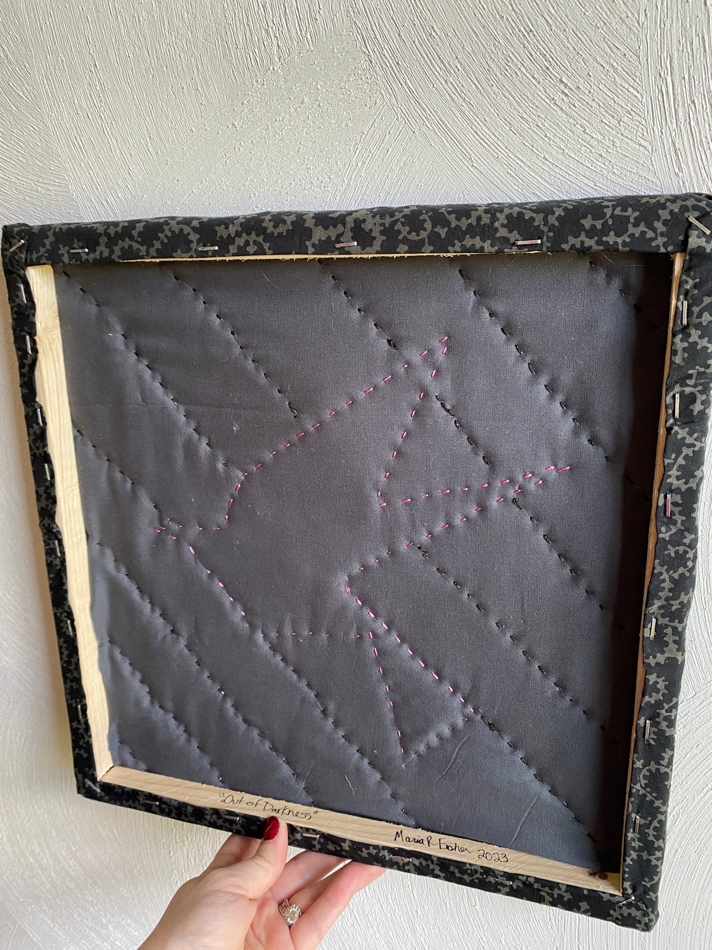 The backside of the framed, quilted wall hanging of a pink bird. It has a black background fabric with 1/2 square triangles framing the picture. It is apart of the "Out of darkness" collection. 
