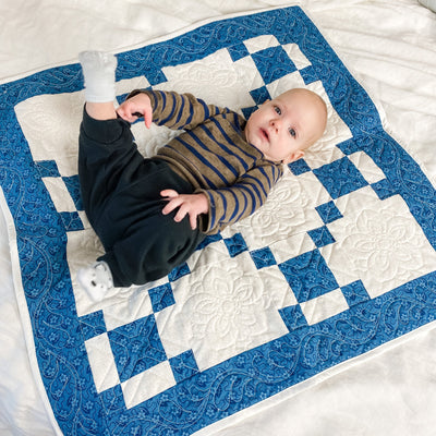 A handmade, heirloom, patchwork of blue nine patch with white background fabric, white backing, 100% cotton batting, Floral top quilting in the white centers, X's in the 9 patch squares, and feathers along the borders, machine top stitched binding, on this baby size quilt