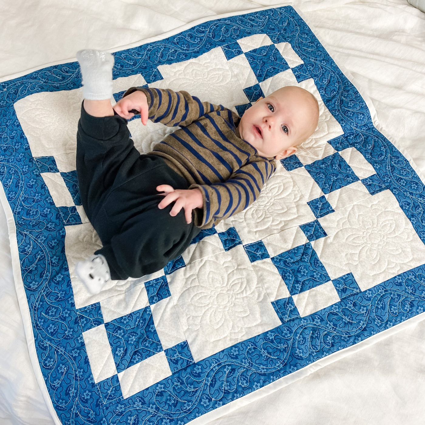 A handmade, heirloom, patchwork of indigo/blue nine patch with white background fabric, natural color backing, 100% cotton batting, Floral top quilting in the white centers, X's in the 9 patch squares, and feathers along the borders, machine top stitched binding, on this baby size quilt