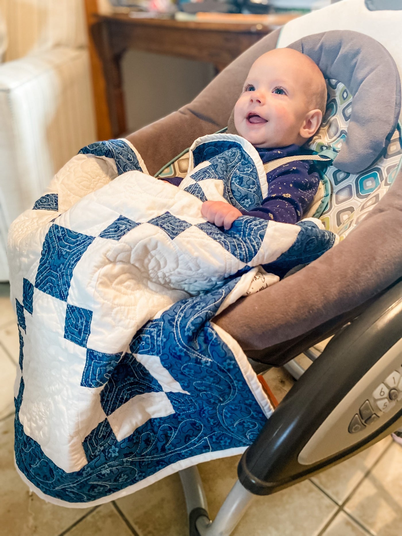 A baby in a swing with a  a handmade, heirloom, patchwork of indigo/blue nine patch with white background fabric, natural color backing, 100% cotton batting, Floral top quilting in the white centers, X's in the 9 patch squares, and feathers along the borders, machine top stitched binding, on this baby size quilt