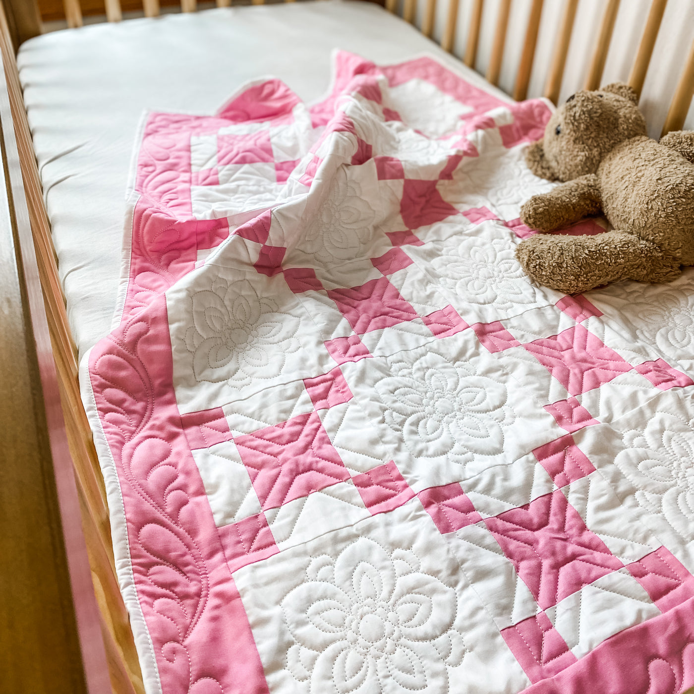 A handmade, heirloom, patchwork of pink nine patch with white background fabric, white backing, 100% cotton batting, Floral top quilting in the white centers, X's in the 9 patch squares, and feathers along the borders, machine top stitched binding, on this crib size quilt