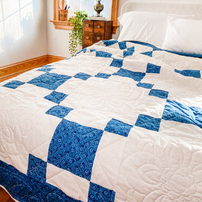 A handmade, heirloom, patchwork of indigo/blue nine patch with white background fabric, natural color backing, 100% cotton batting, Floral top quilting in the white centers, X's in the 9 patch squares, and feathers along the borders, machine top stitched binding, on this queen quilt