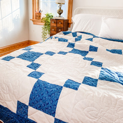 A handmade, heirloom, patchwork of indigo/blue nine patch with white background fabric, natural color backing, 100% cotton batting, Floral top quilting in the white centers, X's in the 9 patch squares, and feathers along the borders, machine top stitched binding, on this king quilt
