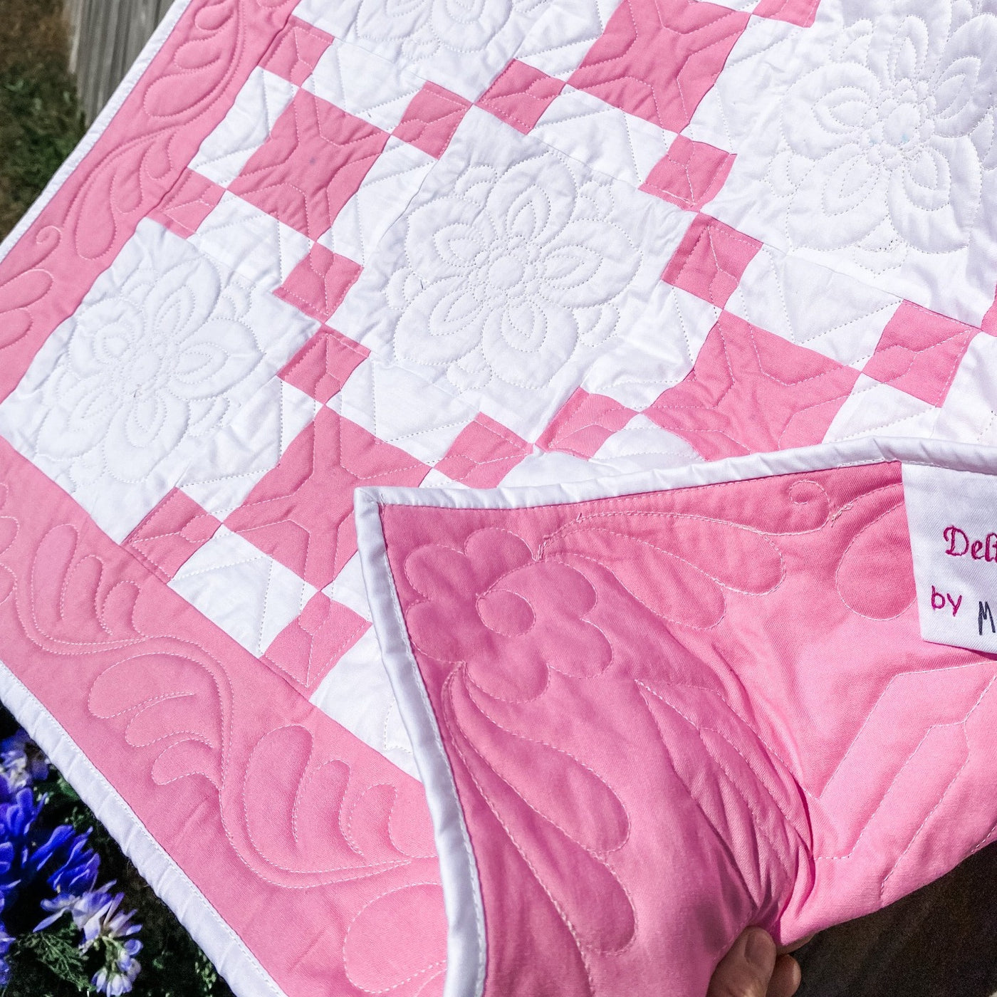 A handmade, heirloom, patchwork of pink nine patch with white background fabric, white backing, 100% cotton batting, Floral top quilting in the white centers, X's in the 9 patch squares, and feathers along the borders, machine top stitched binding, on this crib size quilt