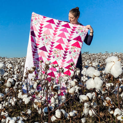 A girl standing in a cotton field holding a handmade, heirloom, patchwork of pink and magenta flying geese on a white background with watercolor bird white backing, straight line top quilting, machine top stitched binding, on this sweet lap size quilt