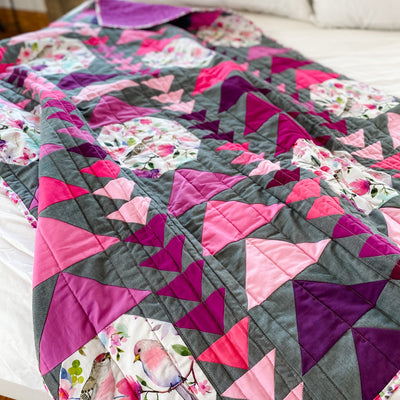 A handmade, heirloom, patchwork of pink and magenta flying geese on a grey/gray background with floral prints, watercolor bird fabric, magenta backing, straight line top quilting, machine top stitched binding, on this lap size quilt