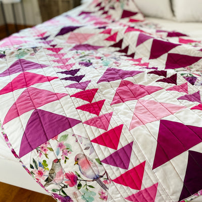 A handmade, heirloom, patchwork of pink and magenta flying geese on a white background with watercolor bird white backing, straight line top quilting, machine top stitched binding, on this sweet lap size quilt