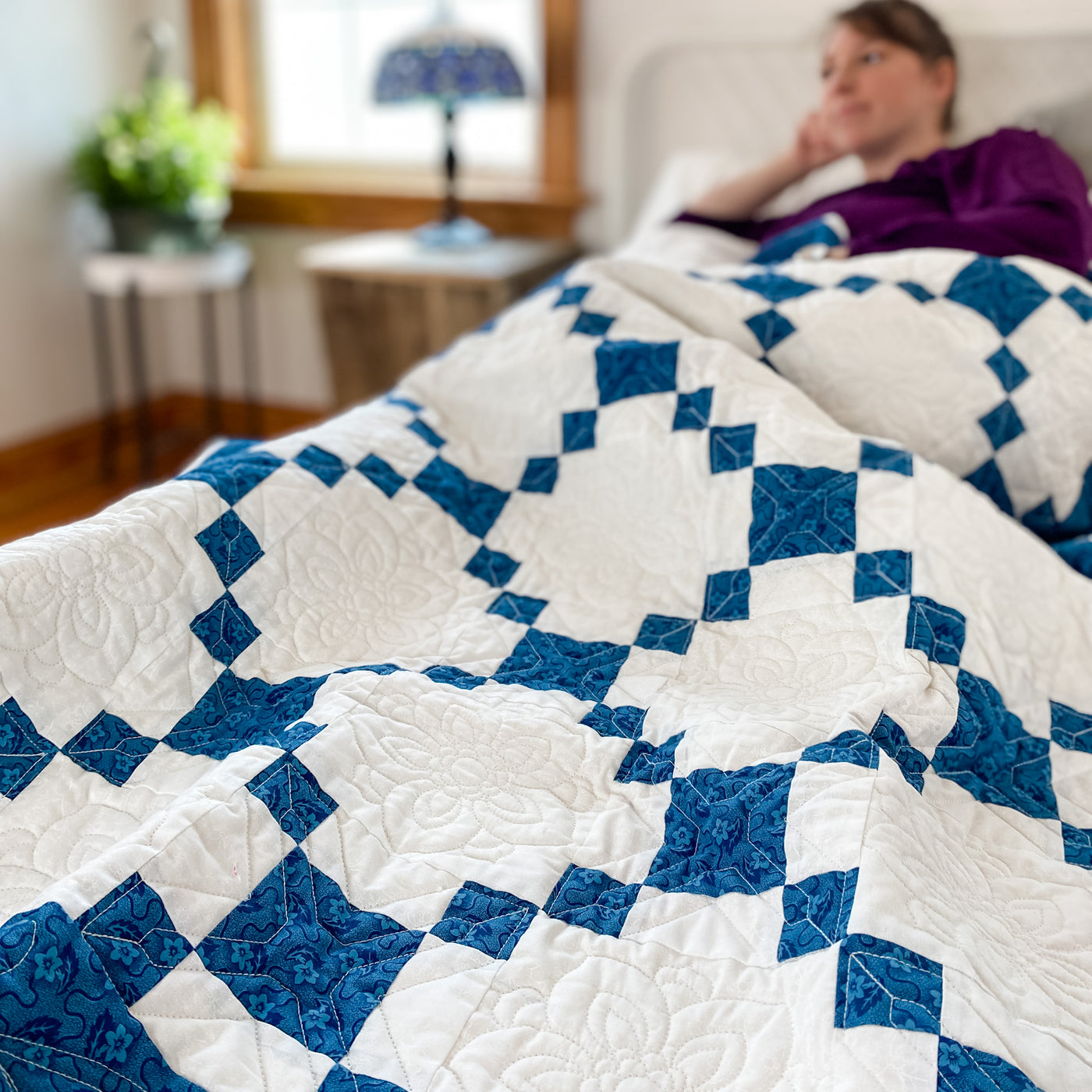 A girl on a bed with a handmade, heirloom, patchwork of indigo/blue nine patch with white background fabric, natural color backing, 100% cotton batting, Floral top quilting in the white centers, X's in the 9 patch squares, and feathers along the borders, machine top stitched binding, on this baby size quilt