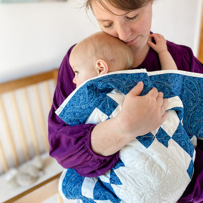 A mom and baby snuggling in a handmade, heirloom, patchwork of indigo/blue nine patch with white background fabric, natural color backing, 100% cotton batting, Floral top quilting in the white centers, X's in the 9 patch squares, and feathers along the borders, machine top stitched binding, on this baby size quilt