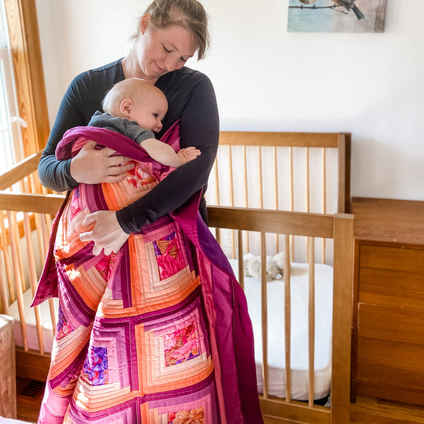 A mom and baby snuggled in A brightly colored purple and orange colored quilt. It is a handmade, patchwork, log cabin style lap quilt