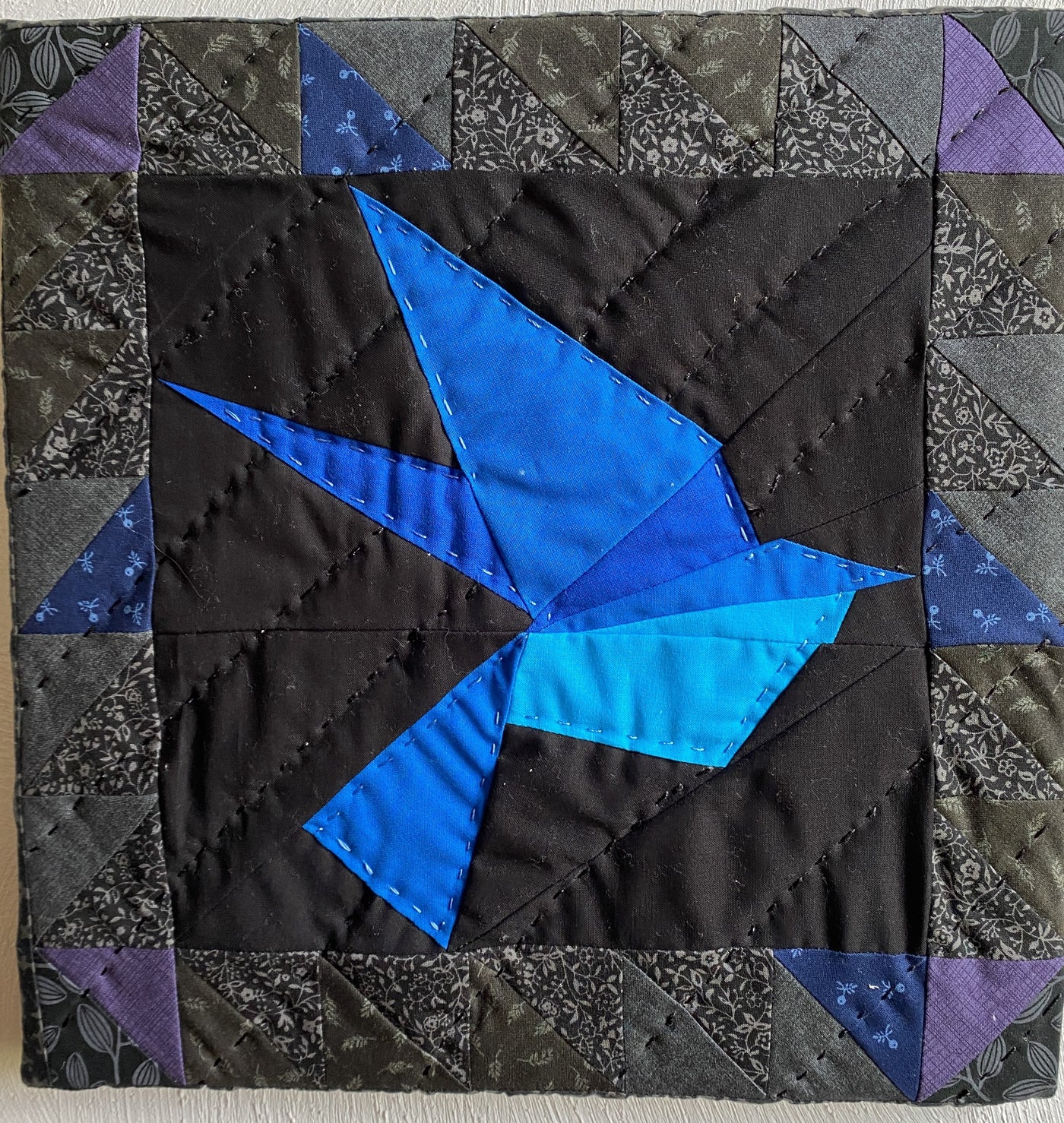 This framed, quilted wall hanging of a blue bird. It has a black background fabric with 1/2 square triangles framing the picture. It is apart of the "Out of darkness" collection. 